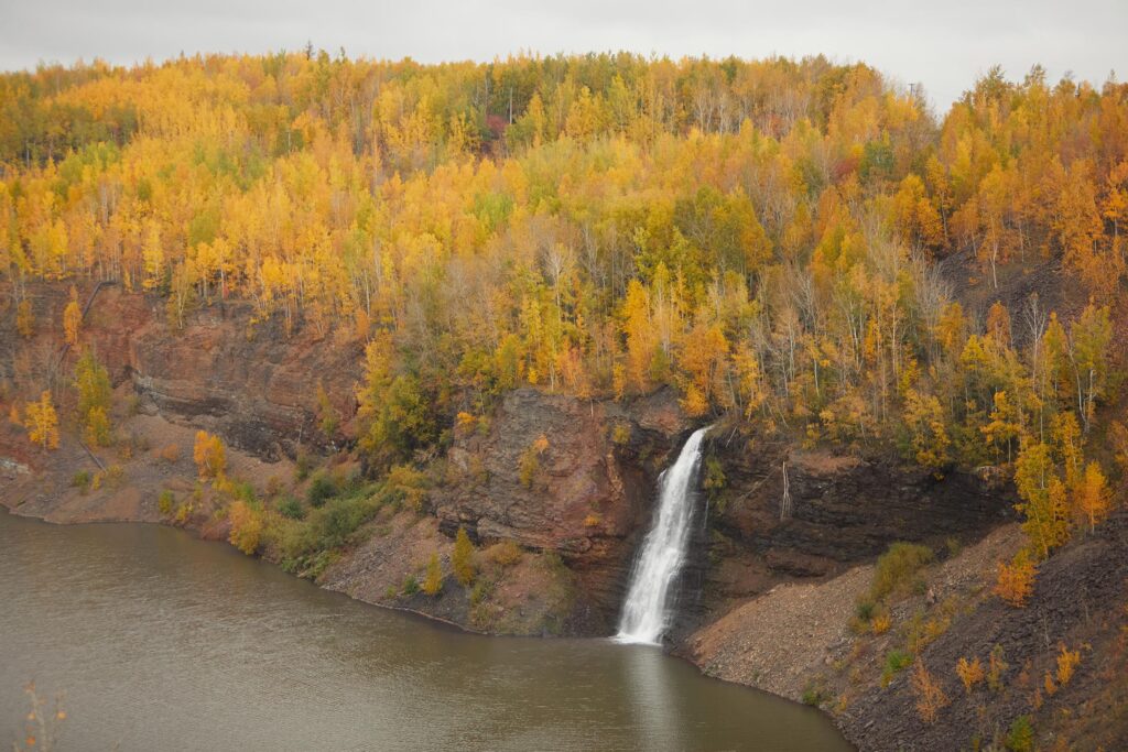 A waterfall protrudes from a forest during fall into an open lake