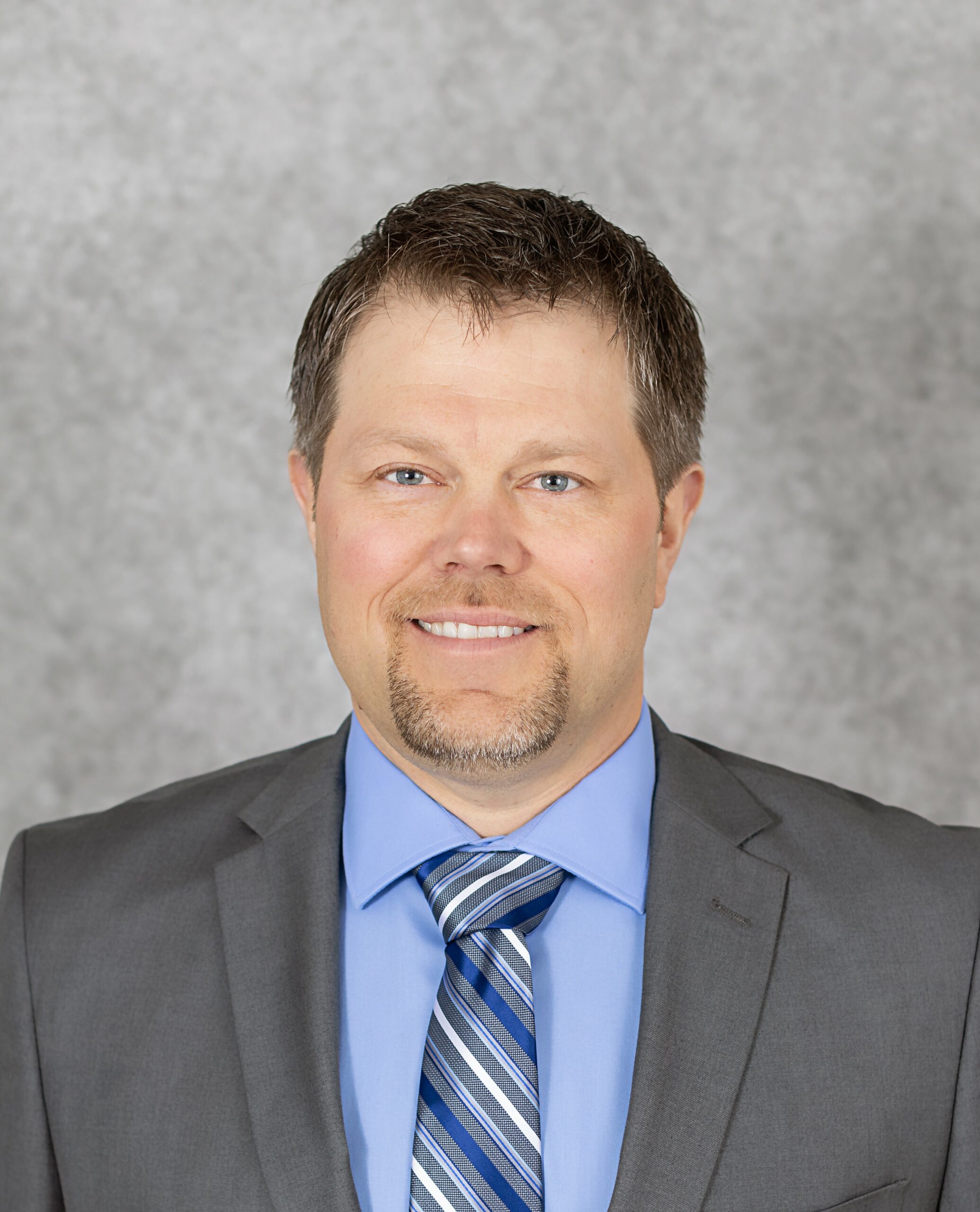 A headshot of a white man with brown hair and goatee in a suit and tie.